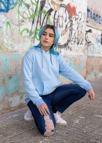 Blue haired Teenage girl in light blue oversize hoodie and looking aside. Blue haired teen girl sitting on her haunches near graffiti wall. Clothing mockup  - Photo, Image