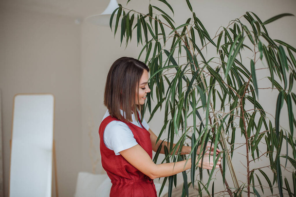 Smiling young brunette woman in a white t-shirt and red overalls is caring for a home tree. Ties the trunk with a brown rope - Zdjęcie, obraz