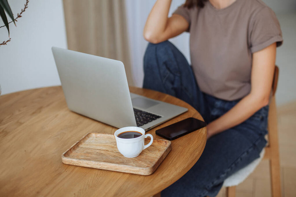 Smiling young brunette woman with short haircut in brown t-shirt and blue jeans working remotely from home on laptop at beige table. Face not visible - Foto, afbeelding