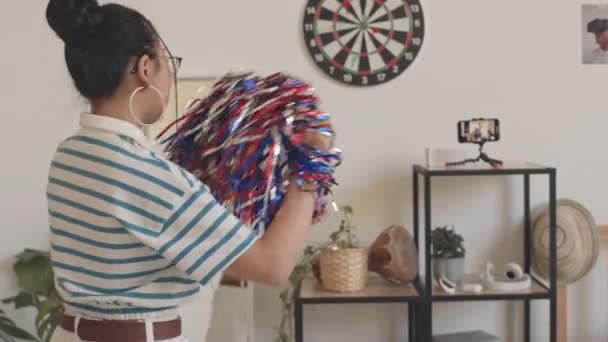 Hátulnézeti felvétel tinédzser pompomlányról csíkos pólóban videofelvétel az okostelefonján, ahogy otthon pomponlányokkal táncol. - Felvétel, videó
