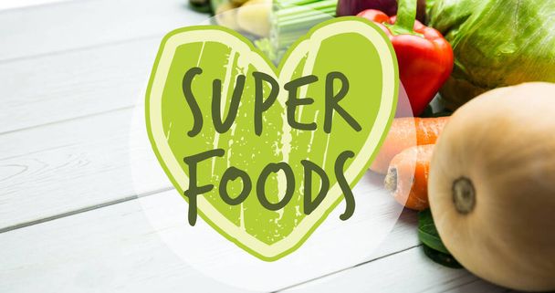 Image of superfoods text in green on green heart, over fresh vegetables on white boards. vegan day, organic vegetable produce and healthy eating concept digitally generated image. - Фото, зображення