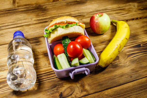 Fles water, appel, banaan en lunchdoos met hamburgers en verse groenten op een houten tafel - Foto, afbeelding