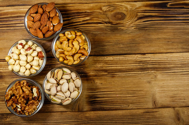 Assortment of nuts on wooden table. Almond, hazelnut, pistachio, walnut and cashew in glass bowls. Top view, copy space. Healthy eating concept - Photo, Image