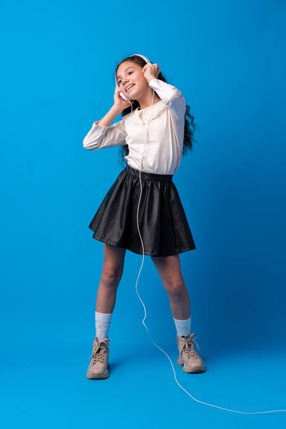 Schoolgirl with headphones listening to music against blue background - Photo, Image