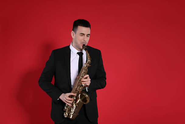 Young man in elegant suit playing saxophone on red background - Fotó, kép