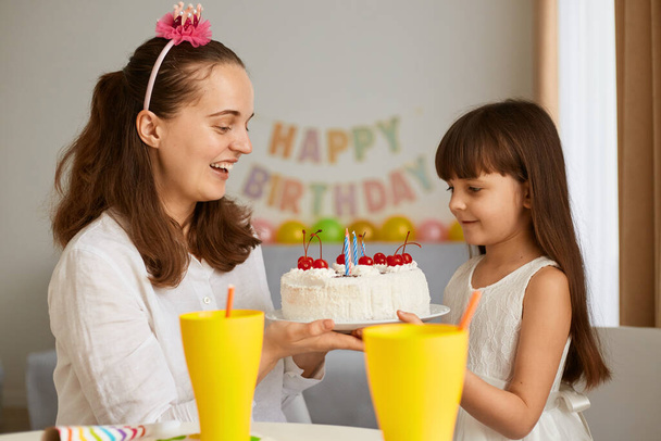 Indoor shot of woman with daughter cerebrating birthday, mother holding cake and congratulating little girl, family posing at home with festive inscription and balloons on background. - Foto, immagini