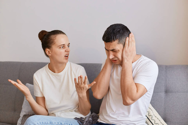 Young couple having conflict, angry fury woman sitting on couch and screaming on her husband with raised arms, having quarrel, problems in relationship. - Fotó, kép