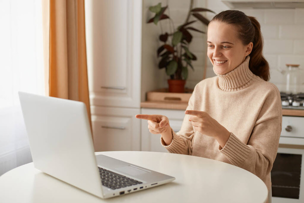 Portrait of attractive positive woman wearing beige sweater sitting in kitchen and working online on laptop, pointing at notebook display, expressing happiness. - Foto, afbeelding