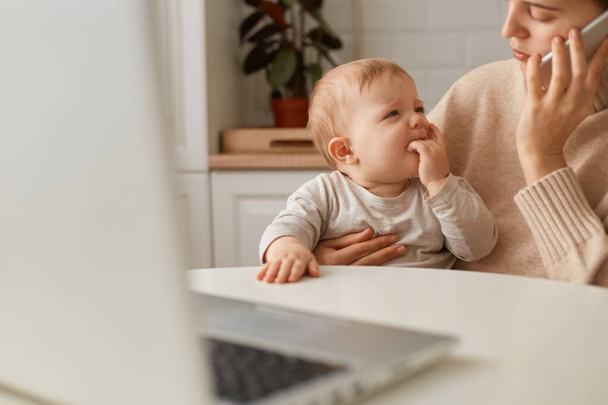 Cropped image woman wearing beige sweater sitting in kitchen and working online on laptop and talking via smartphone, looking after baby girl, calming down crying daughter. - Φωτογραφία, εικόνα