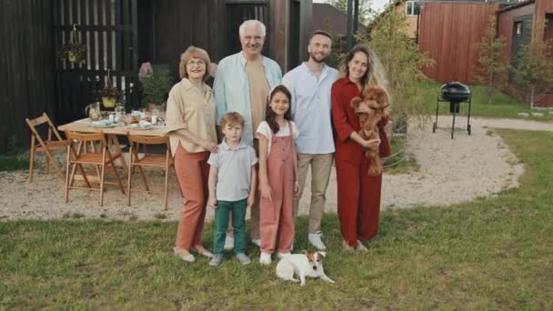 Довгий знімок великої кавказької сім'ї з трьох поколінь і двох собак, що сидять перед подвір'ям літнього будинку в теплий день, посміхаючись і дивлячись на камеру - Кадри, відео