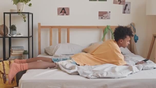 PAN toma de gen z Biracial adolescente saltando abajo en su cama con teléfono inteligente - Imágenes, Vídeo