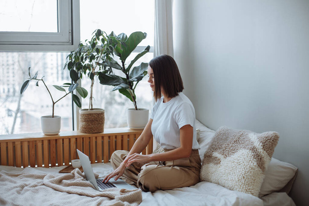 Smiling young brunette woman with a short haircut in a white t-shirt and beige trousers sits on the bed at home and works on a laptop. On the bed is a wooden tray with a white mug of coffee - Photo, image