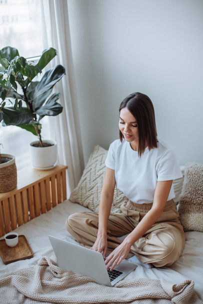 Smiling young brunette woman with a short haircut in a white t-shirt and beige trousers sits on the bed at home and works on a laptop. On the bed is a wooden tray with a white mug of coffee - Фото, зображення