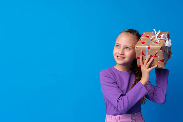 Happy little girl with long hair holding a gift box on a blue background - Foto, imagen