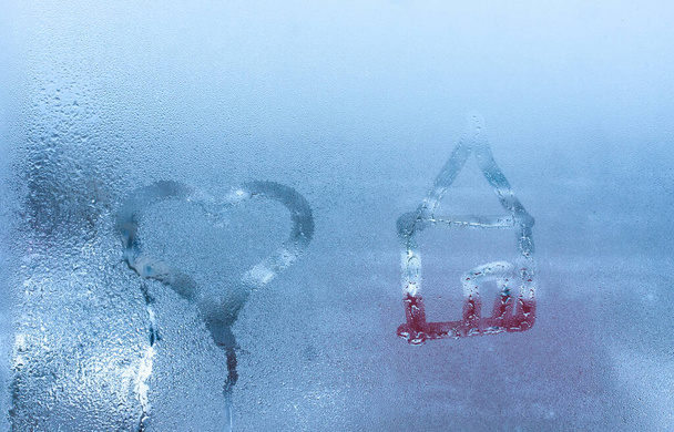 Közelkép a szerelmi szívről és az otthoni kondenzációról, ami az izzadt ablaküvegre van írva. A kéz rajzol egy szívet és egy házat egy ködös ablakon. - Fotó, kép