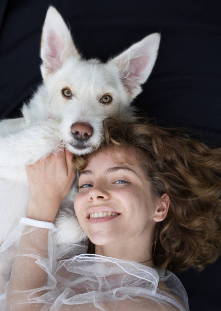 retrato de una linda chica - una adolescente de 17-18 años de edad junto a su perro blanco sobre un fondo oscuro. la similitud y amistad entre la propietaria y la mascota. Día mundial del perro - Foto, imagen