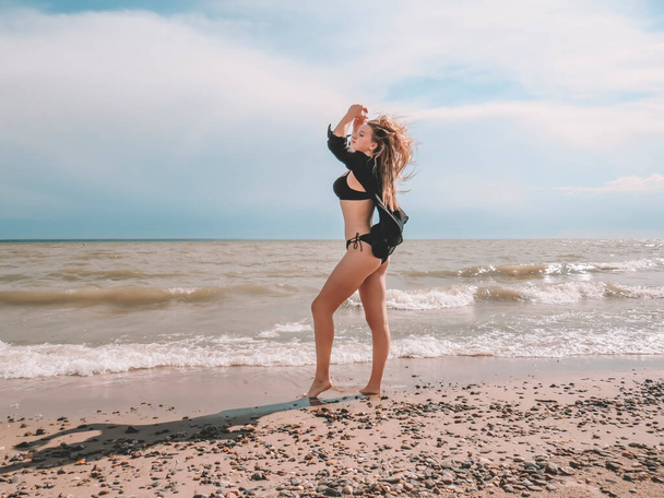 A blonde young woman in a black open shirt and a black bikini swimsuit with a beautiful body shape dances on a sandy beach against the background of sea waves on a windy summer day - Fotó, kép