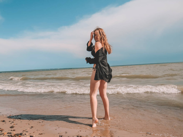 A blonde young woman in a black open shirt and a black bikini swimsuit with a beautiful body shape dances on a sandy beach against the background of sea waves on a windy summer day - Fotoğraf, Görsel