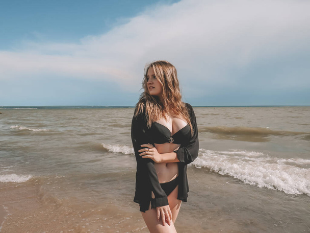 A young beautiful woman with loose blonde hair in a black bikini and an open short shirt against the background of sea waves on a sandy beach on a hot summer day. The concept of love, joy, happiness, freedom, outdoor recreation, active lifestyle - 写真・画像