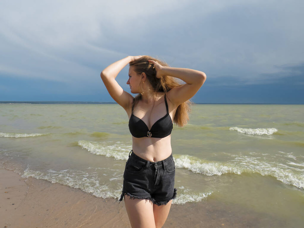 A blonde young woman in black shorts and a black bra on a beautiful chest with a beautiful slender figure dances on a sandy beach against the background of sea waves and a dark blue sky on a windy summer day before an approaching thunderstorm - Foto, imagen