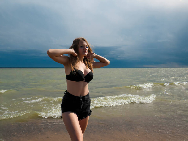 A blonde young woman in black shorts and a black bra on a beautiful chest with a beautiful slender figure dances on a sandy beach against the background of sea waves and a dark blue sky on a windy summer day before an approaching thunderstorm - Foto, afbeelding