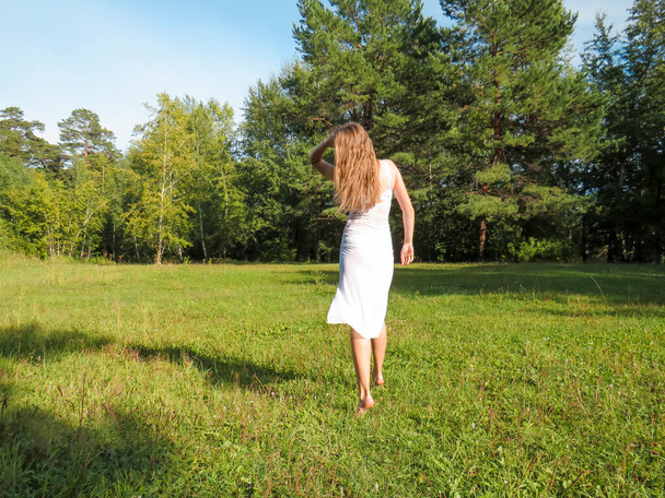 Barefoot red-haired young woman with a slender figure in a white silk dress having fun and enjoying the freedom of a sunny forest glade on a hot summer day, the concept of walking and relaxing in nature, active lifestyle - Fotó, kép