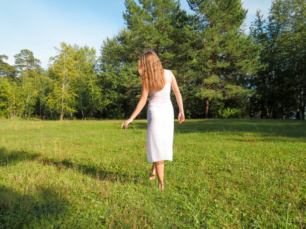 Barefoot red-haired young woman with a slender figure in a white silk dress having fun and enjoying the freedom of a sunny forest glade on a hot summer day, the concept of walking and relaxing in nature, active lifestyle - Foto, imagen
