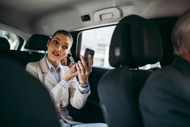 Good looking young business woman sitting on backseat in luxury car and fixing her makeup. Transportation in corporate business concept. - Фото, зображення