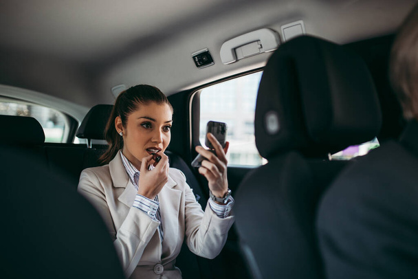 Good looking young business woman sitting on backseat in luxury car and fixing her makeup. Transportation in corporate business concept. - Foto, imagen