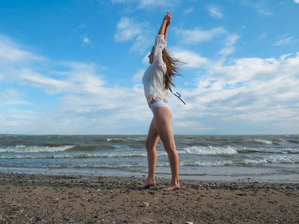 A young blonde woman with disheveled hair in a white shirt and a bikini with slender tanned legs on the beach on the wet sand against the background of sea waves and blue cloudy sky on a hot windy summer day, the concept of an active lifestyle - 写真・画像