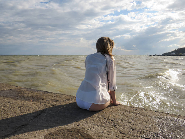 young blonde woman with long hair disheveled in the wind in a white shirt and a white bikini with long slender tanned legs, sitting on the edge of a stone pier against the background of foamy sea waves and blue sky in the sunlight on windy summer day - 写真・画像