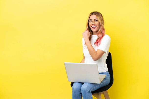 Young woman sitting on a chair with laptop over isolated yellow background celebrating a victory - Photo, image