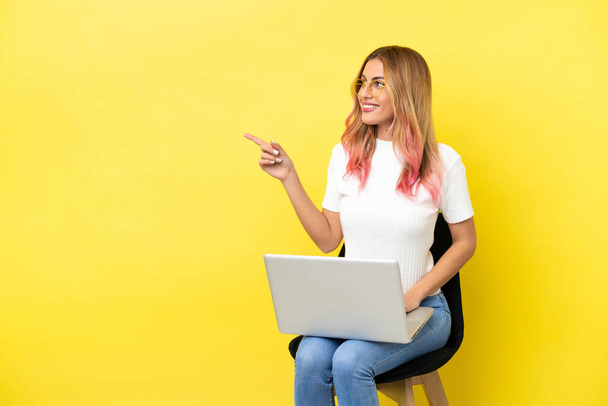 Dizüstü bilgisayarlı, sarı arka planda oturan genç bir kadın çözümü bulmak için parmağını kaldırıyor. - Fotoğraf, Görsel