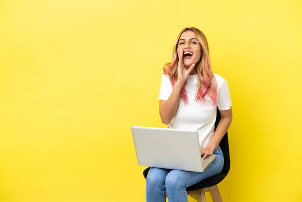 Young woman sitting on a chair with laptop over isolated yellow background shouting with mouth wide open - Фото, изображение