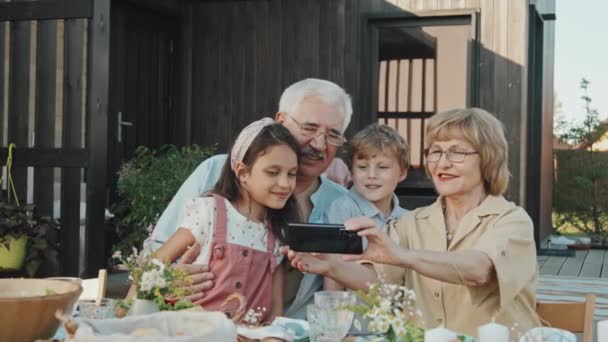 Pas-up dospělé bělošky držící smartphone, mluví selfie se svými malými vnoučaty a manželem, sedí u stolu, mluví a usmívá se před letním domem za slunečného dne - Záběry, video