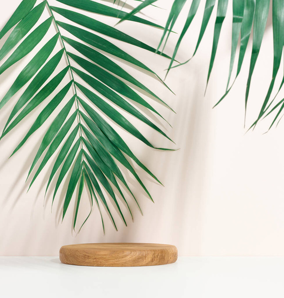 stage for displaying products, cosmetics with a round wooden podium and a green palm leaf. Shadow on the background - Foto, afbeelding