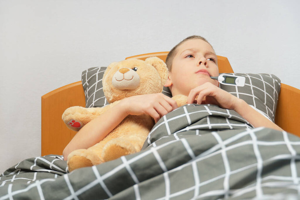 un niño enfermo triste con un termómetro en la boca yace en la cama con un oso de juguete. niño enfermo en casa - Foto, Imagen