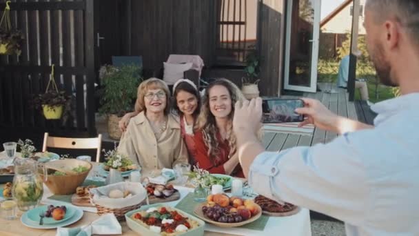Olkapään yli mies käyttää älypuhelinta ottamaan kuvia naispuolisista valkoihoisista perheenjäsenistä, jotka istuvat pöydässä kesämökin ulkopuolella lämpimänä aurinkoisena päivänä hymyillen - Materiaali, video