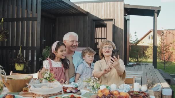 Aufstehen einer gestandenen kaukasischen Frau, die ihr Smartphone in der Hand hält, Selfie mit ihren kleinen Enkeln und ihrem Mann macht und an einem sonnigen Tag am Tisch vor dem Sommerhaus sitzt - Filmmaterial, Video