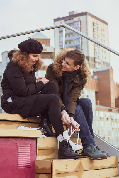 A young couple in love is going ice skating. A young man helps his girlfriend to put on skates on the podium of a skating rink in the city. - 写真・画像
