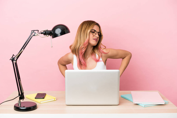 Mujer joven estudiante en un lugar de trabajo con una computadora portátil sobre fondo rosa que sufre de dolor de espalda por haber hecho un esfuerzo - Foto, Imagen