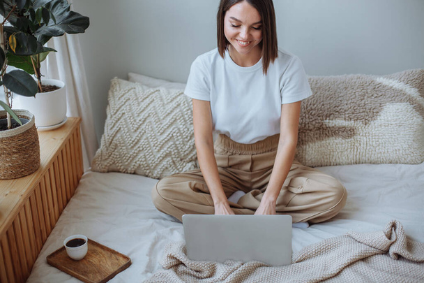 Smiling young brunette woman with a short haircut in a white t-shirt and beige trousers sits on the bed at home and works on a laptop. On the bed is a wooden tray with a white mug of coffee - Foto, afbeelding