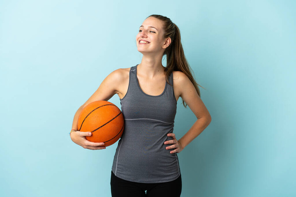Mujer joven jugando baloncesto aislado sobre fondo azul posando con los brazos en la cadera y sonriendo - Foto, Imagen
