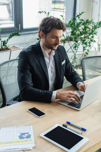 businessman in stylish formal wear typing on laptop near devices on desk - Foto, imagen