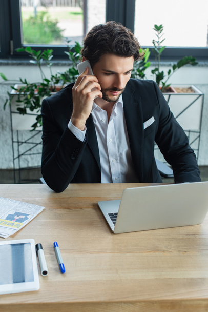 businessman in black blazer talking on smartphone near laptop at workplace - Фото, зображення