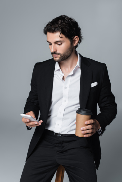 stylish brunette man with coffee to go using mobile phone while sitting isolated on grey - Φωτογραφία, εικόνα