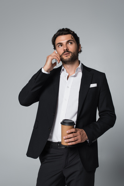 brunette man in black blazer holding coffee to go and talking on smartphone isolated on grey - Fotografie, Obrázek