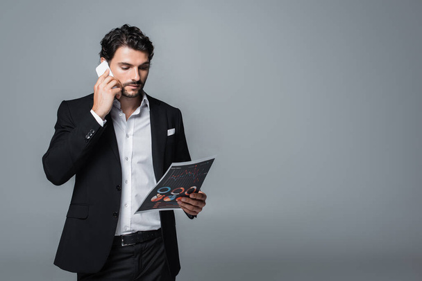 hombre de negocios en ropa formal mirando gráficos y hablando en el teléfono móvil aislado en gris - Foto, Imagen