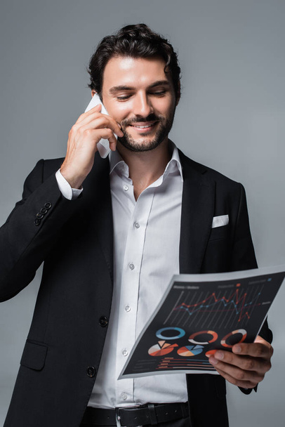 homem de negócios sorrindo olhando para infográficos e falando no celular isolado no cinza - Foto, Imagem