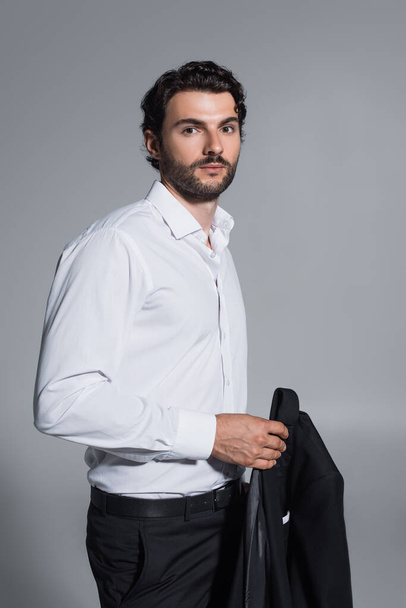 brunette man in white shirt holding black blazer while looking at camera isolated on grey - Foto, immagini